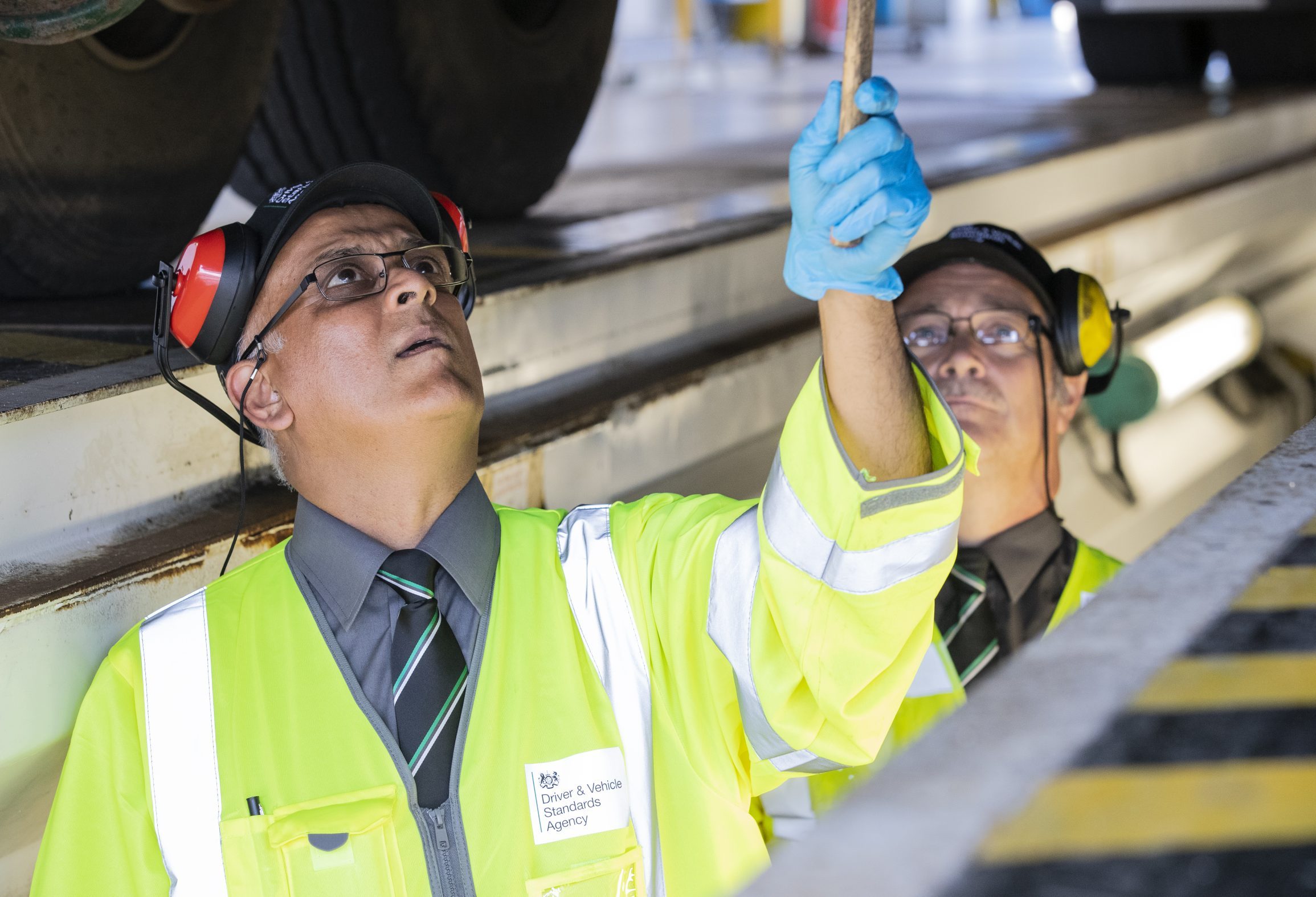 Two DfT Engineers in high-visibility jackets, standing beneath a vehicle and using tools to inspect an issue.